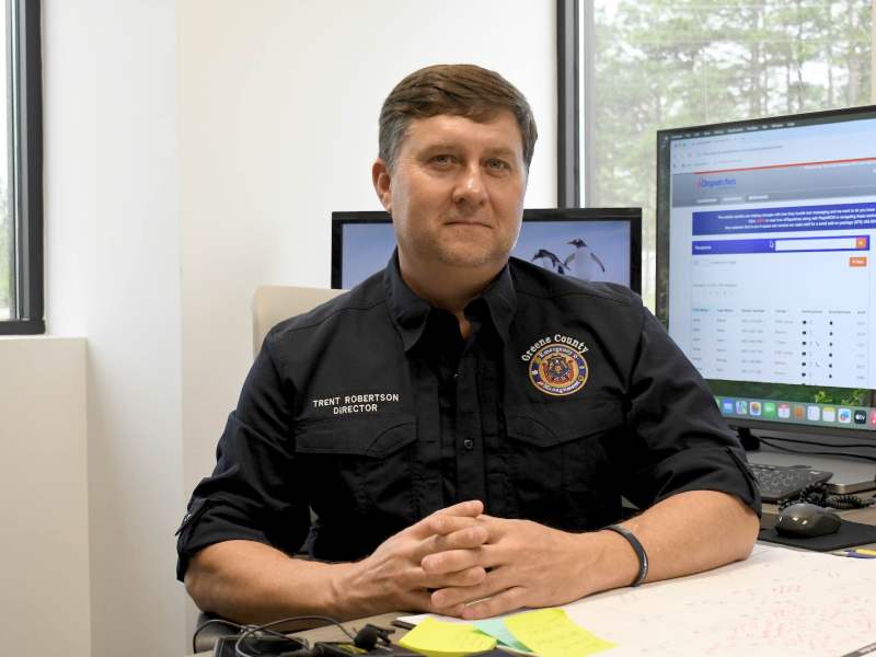 Trent Robertson sits at a desk and smiles at the camera. His shirt reads “Trent Robertson, Director” and displays a Greene County Emergency Management logo. Two computer monitors are behind him.