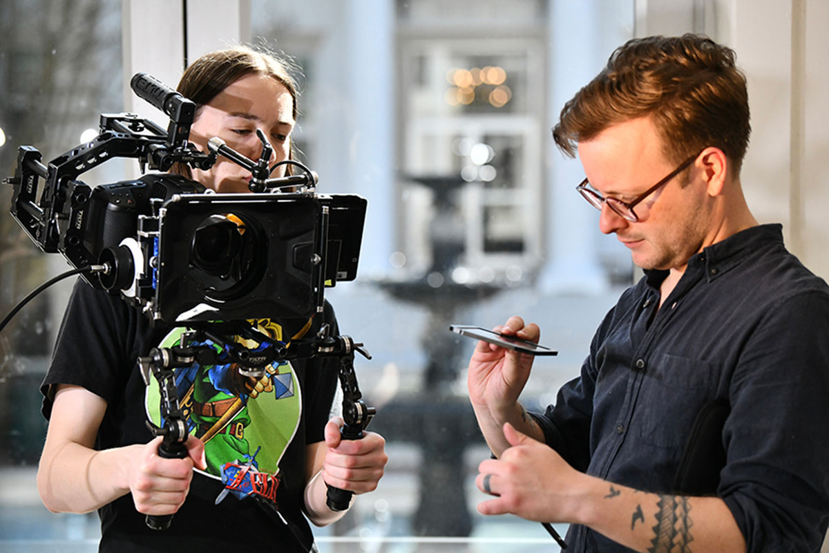 Two people looking down, the one on the left is holding a shoulder mounted camera rig and the other is holding something up