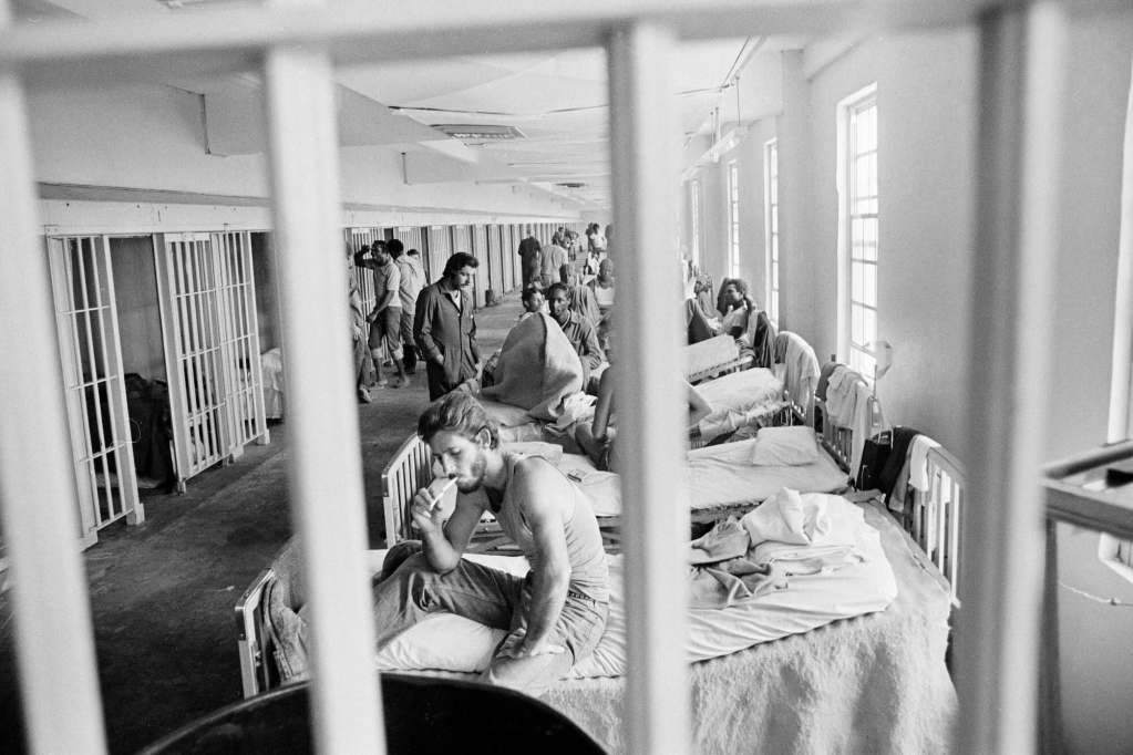 An older black and white photo of men inside a prison hallway filled with beds behind bars