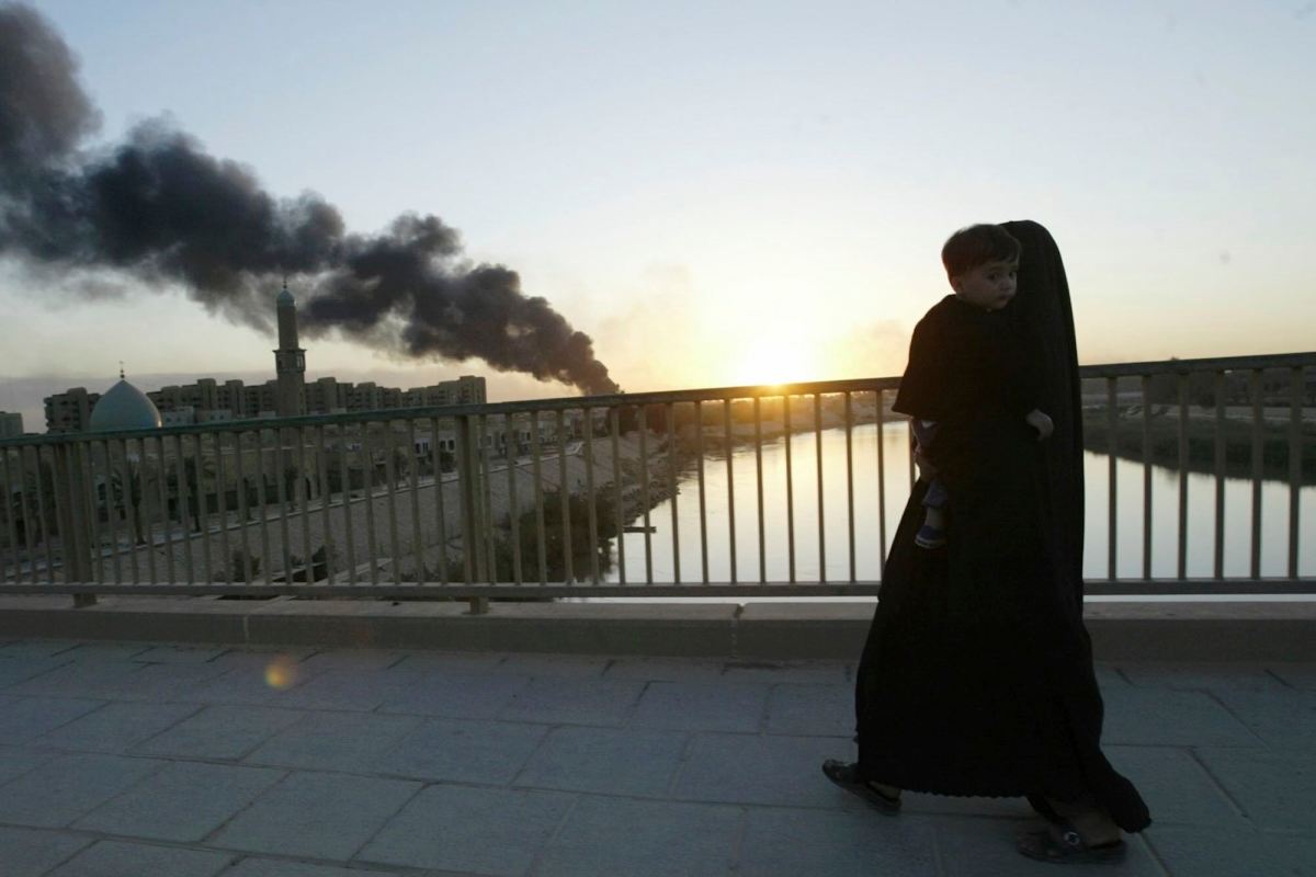 A woman wearing a burqa carrying a young child on a bridge. A plume of dark smoke rises in the sky in the background.