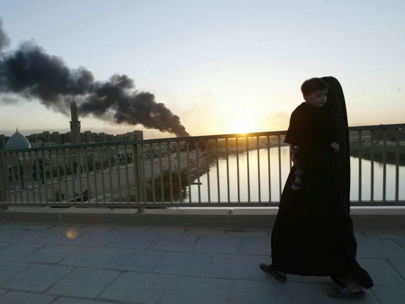 A woman wearing a burqa carrying a young child on a bridge. A plume of dark smoke rises in the sky in the background.