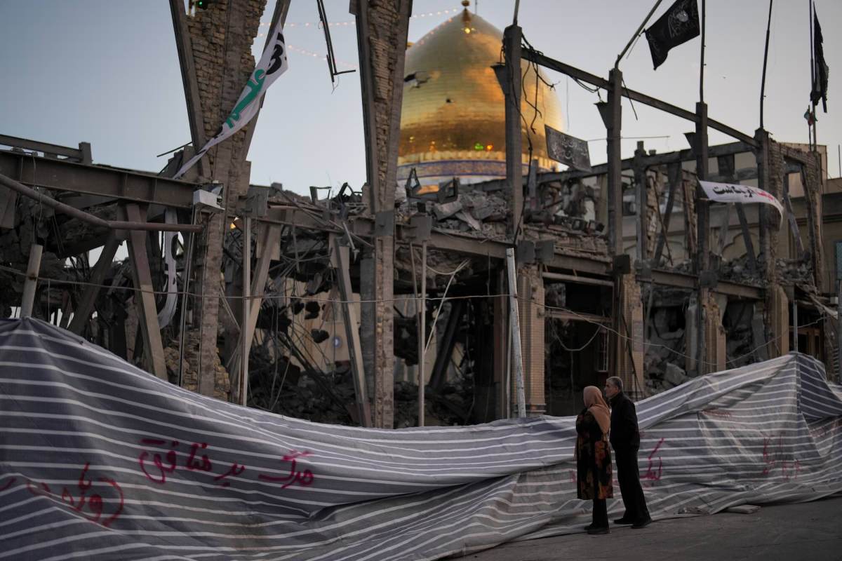 Two people looking at a destroyed building. The golden dome of a mosque is in the background.