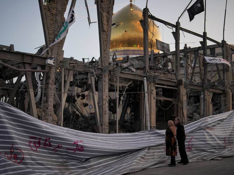 Two people looking at a destroyed building. The golden dome of a mosque is in the background.