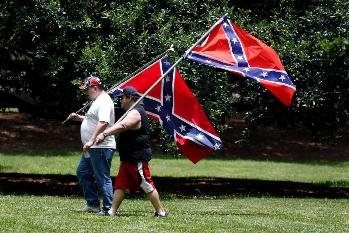Two men in casual clothes carry large confederate flags on poles over their shoulders across a green lawn