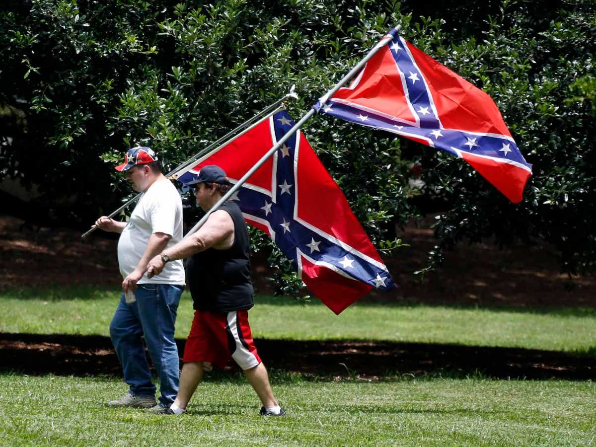 Two men in casual clothes carry large confederate flags on poles over their shoulders across a green lawn