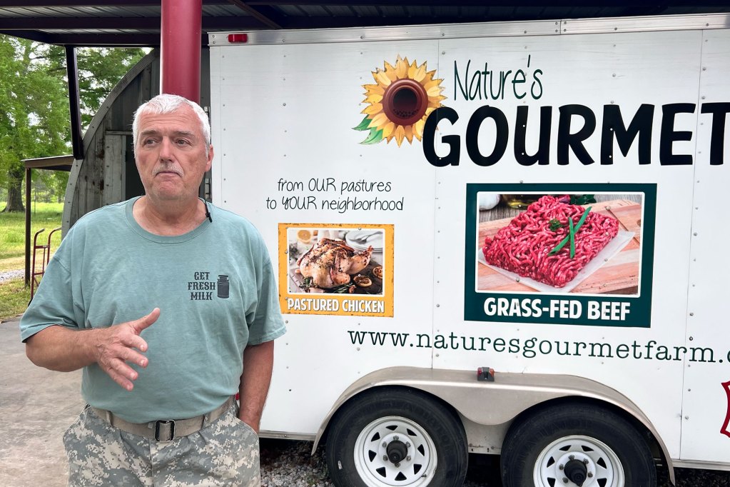 A man in a green shirt and camouflage pants stands next to a Nature's Gourmet Farm trailer.