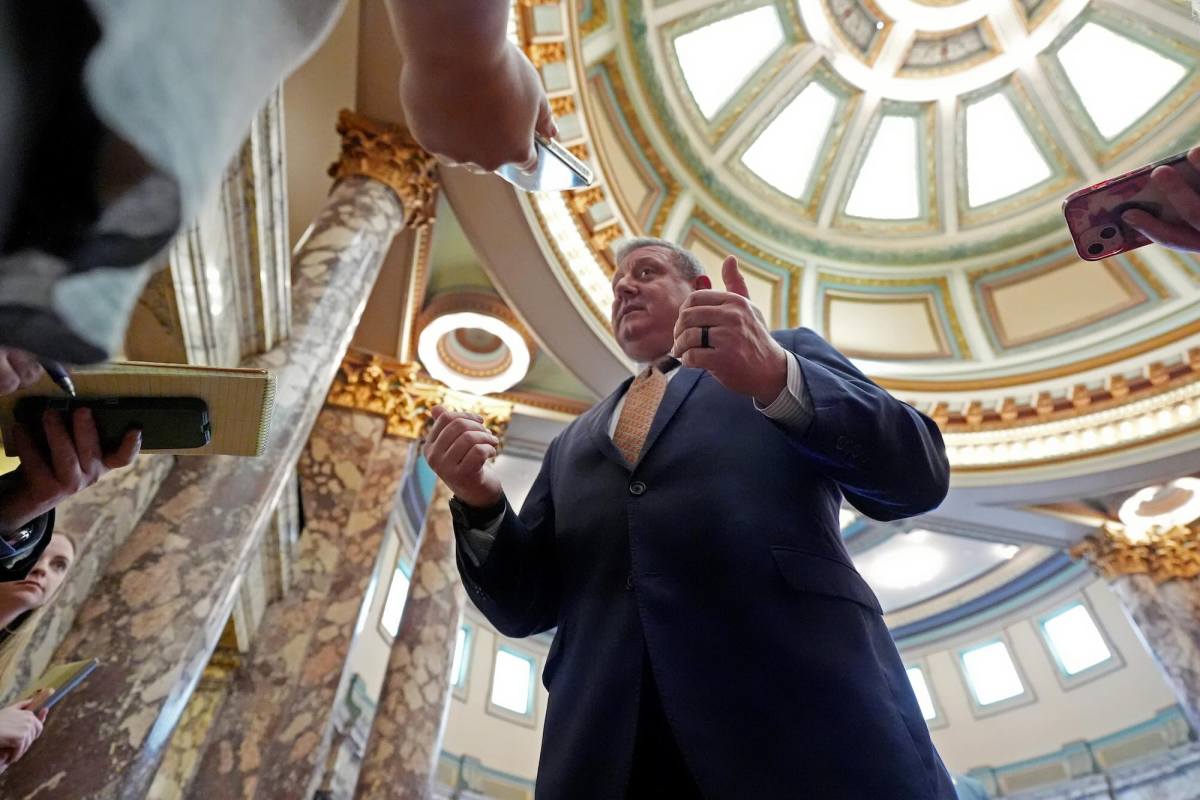 A man in a suit with gold tie speaks to reporters in a marble room with a circular atrium
