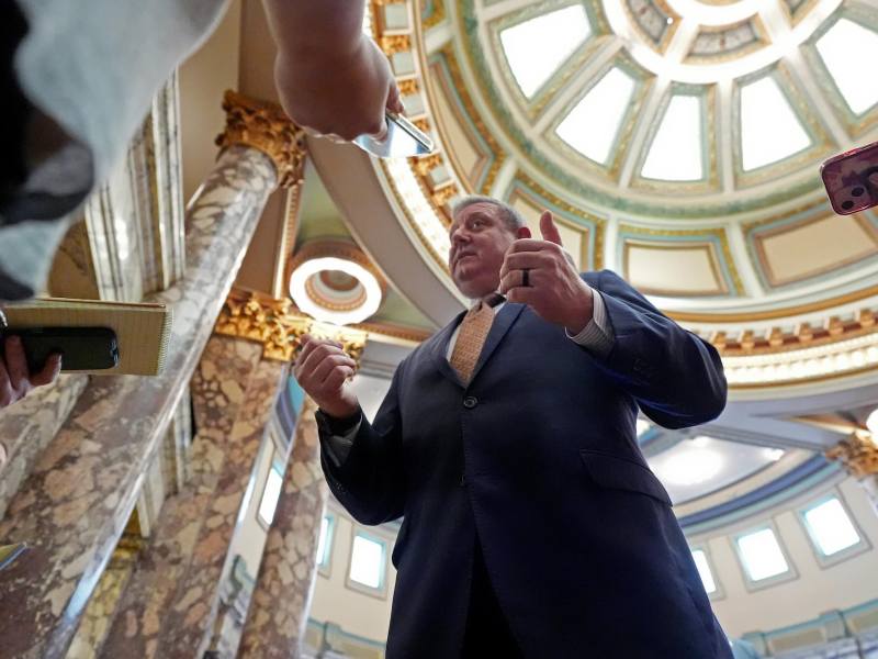 A man in a suit with gold tie speaks to reporters in a marble room with a circular atrium