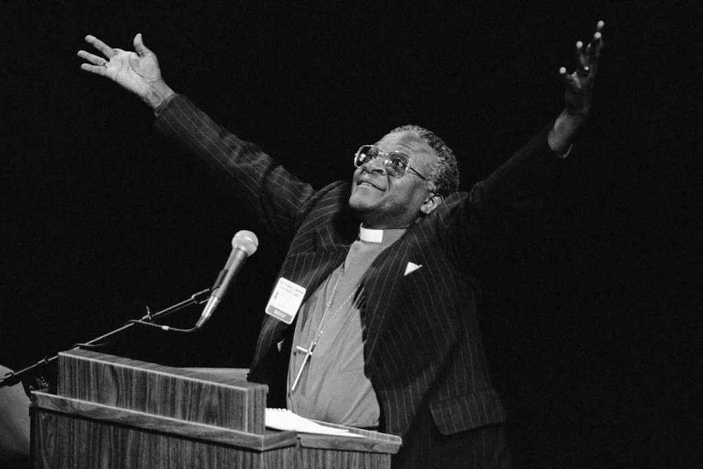 Desmond Tutu in a pinstriped suit jacket and bishop collar stands behind a mic, arms spread wide