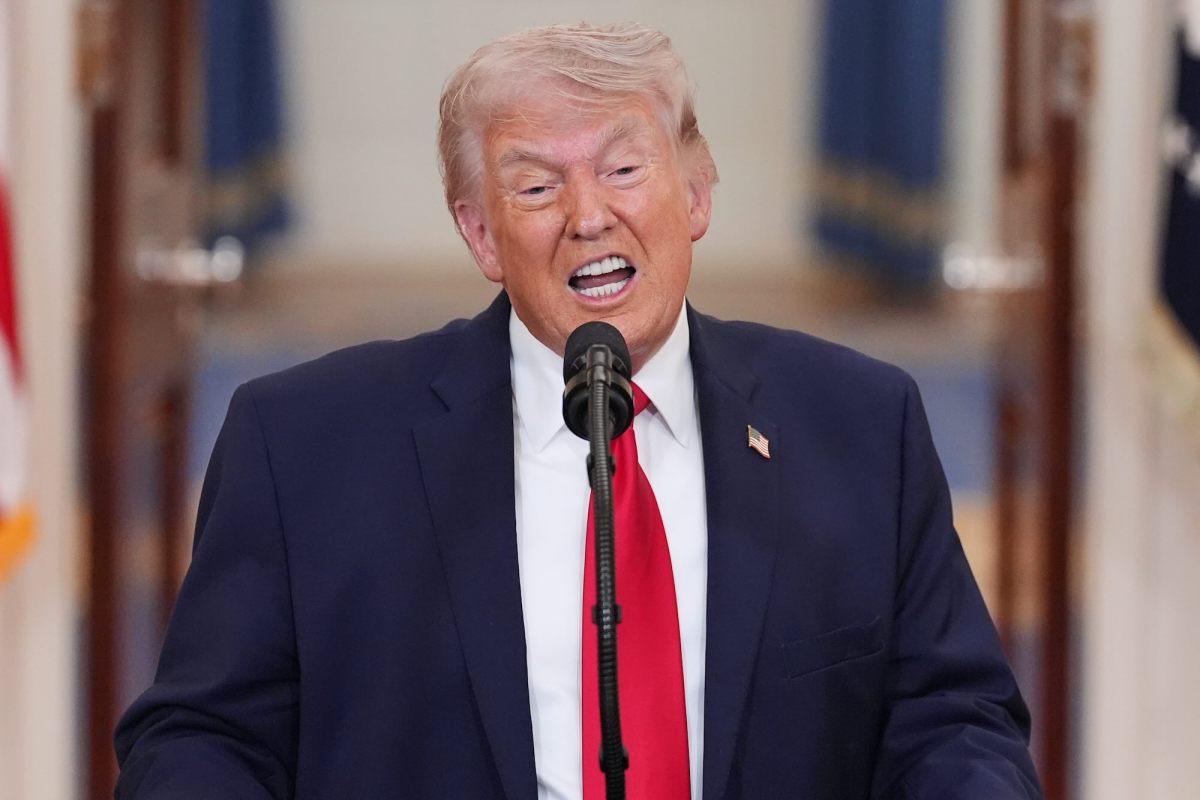 Donald Trump, in a dark navy suit and red tie, speaks at a mic