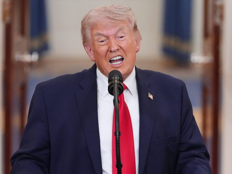 Donald Trump, in a dark navy suit and red tie, speaks at a mic