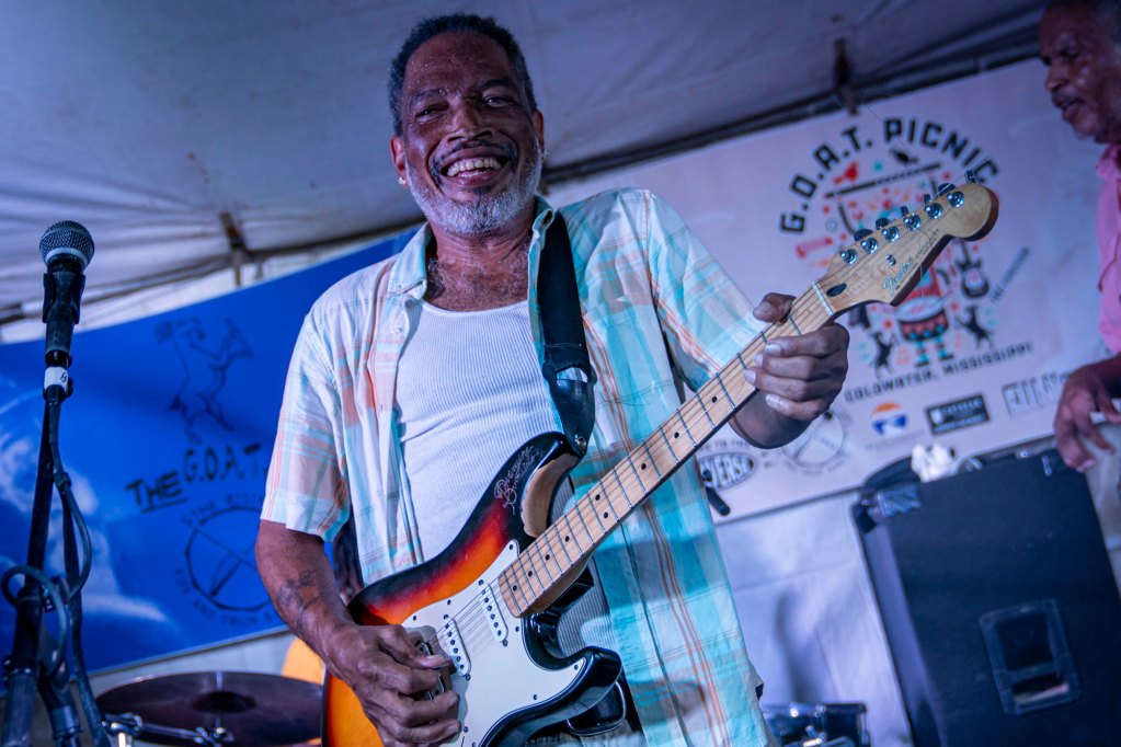 A man plays guitar on stage, a sign behind him reads "GOAT Picnic"
