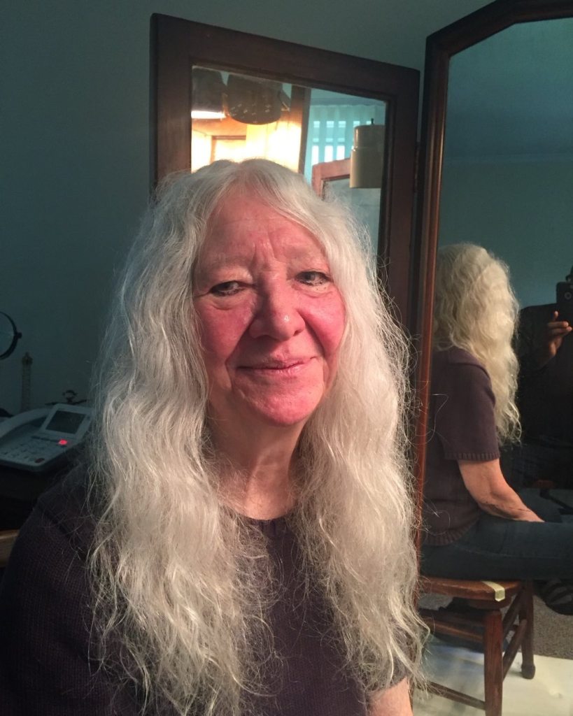 A woman with long white hair sits in a room in front of a mirror