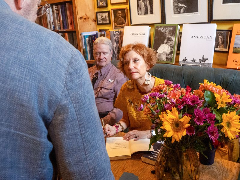 A woman with ginger hair looks intently at a man wearing a grey jacket, whose back is two us, while she signs a book. Another man sits next to her, glancing down at the table.