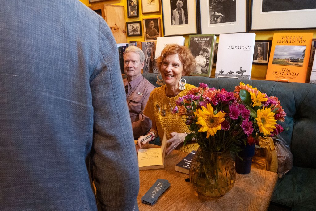A woman with ginger hair looks intently at a man wearing a grey jacket, whose back is two us, while she signs a book. Another man sits next to her, glancing down at the table.