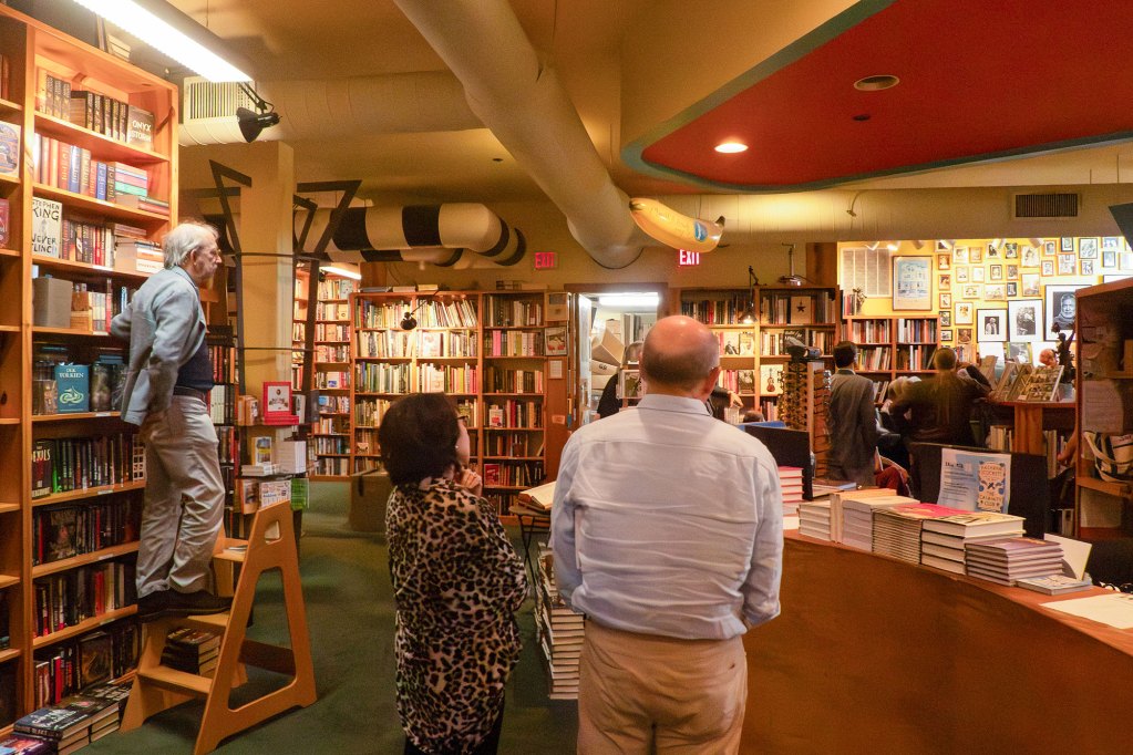 A variety of people stand around a bookstore, looking towards an unseen corner where two women are speaking. One man, on the left, is standing on a 3-step book ladder.