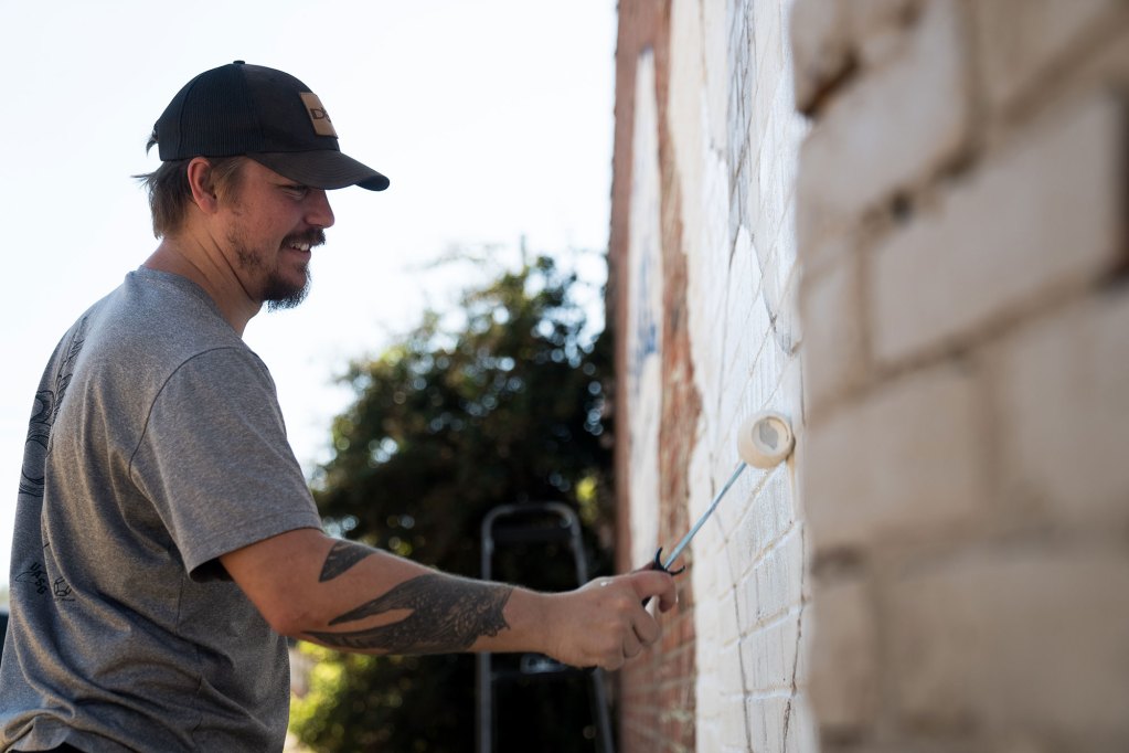 A man is seen using a roller to apply white paint to a brick wall outside