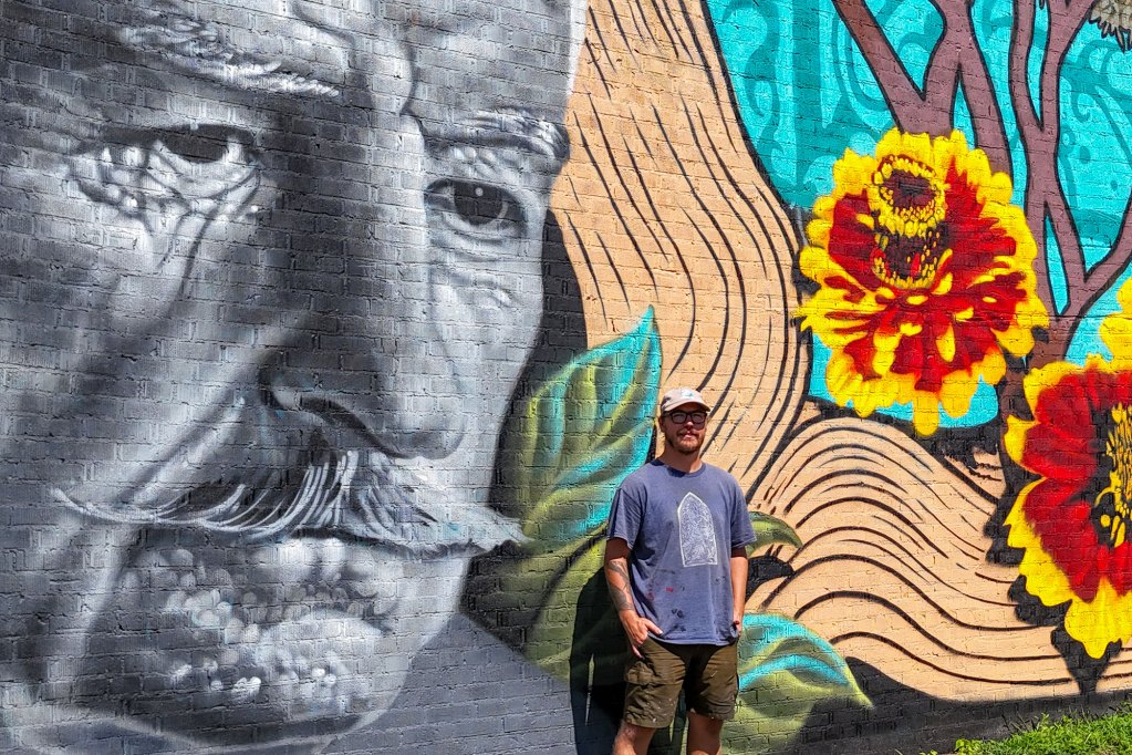 A man stands in front of a large wall mural he painted depicting a man and greenery
