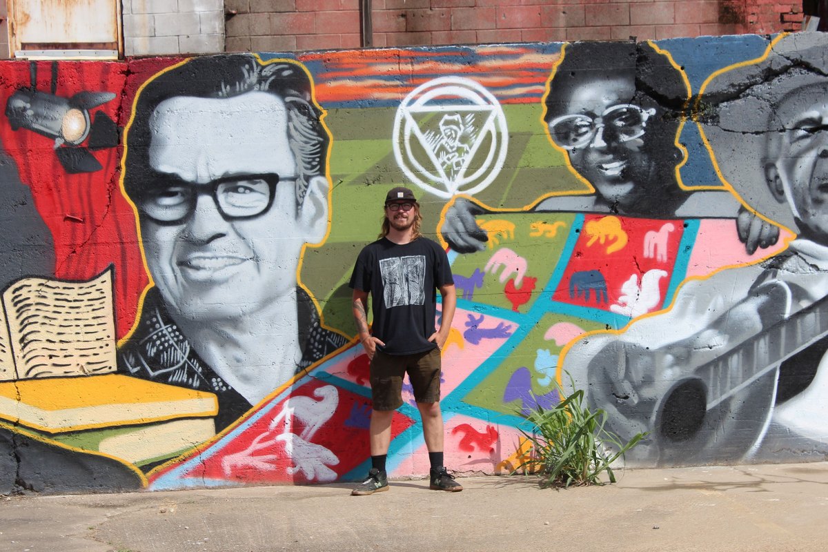 A man stands in front of the colorful wall mural he painted