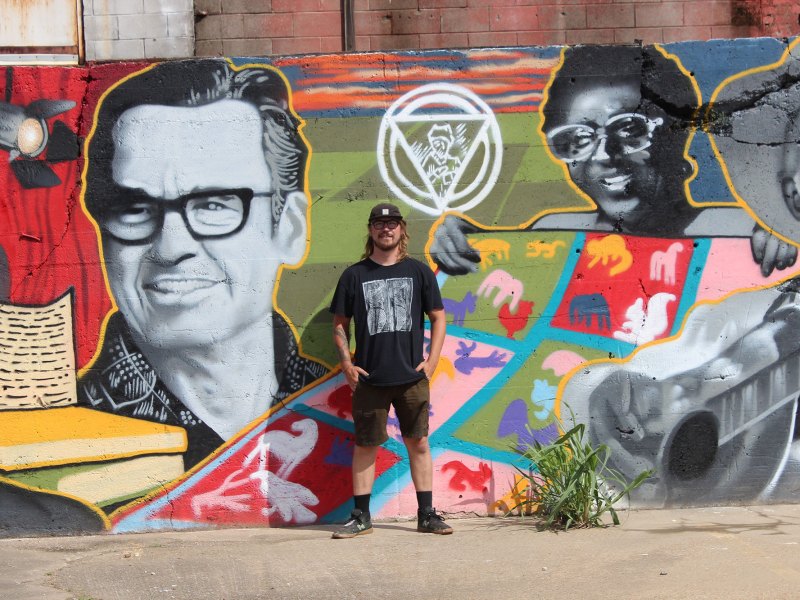 A man stands in front of the colorful wall mural he painted