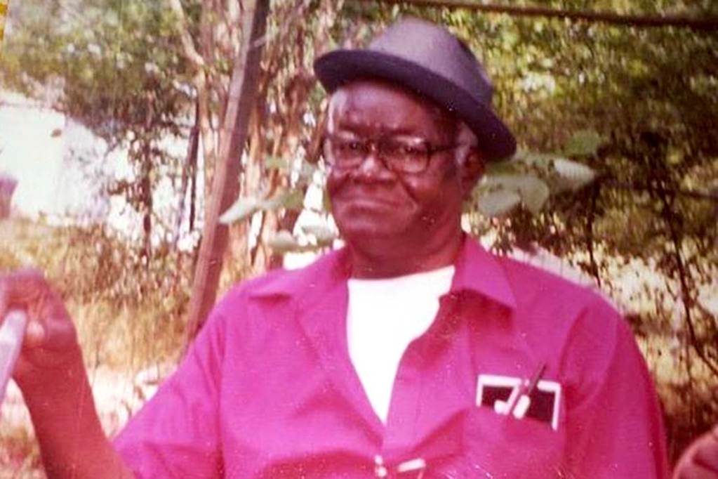 A man in a red shirt and black hat stands outside in the yard