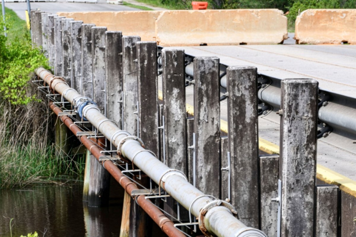 Close-up view of the side of a small bridge showing weathered wooden pilings, metal rail supports and large utility pipes running alongside the roadway above dark water and marsh grass.