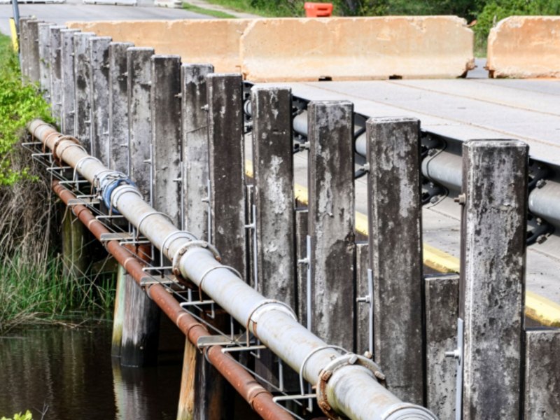 Close-up view of the side of a small bridge showing weathered wooden pilings, metal rail supports and large utility pipes running alongside the roadway above dark water and marsh grass.