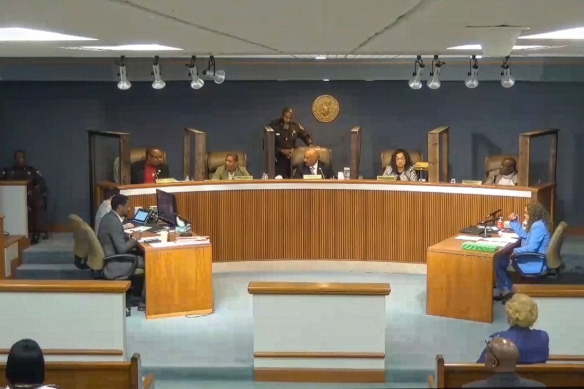 A man in a brown sheriff uniform walks behind a desk that seats two women and three men, facing two occupied desks and an audience.