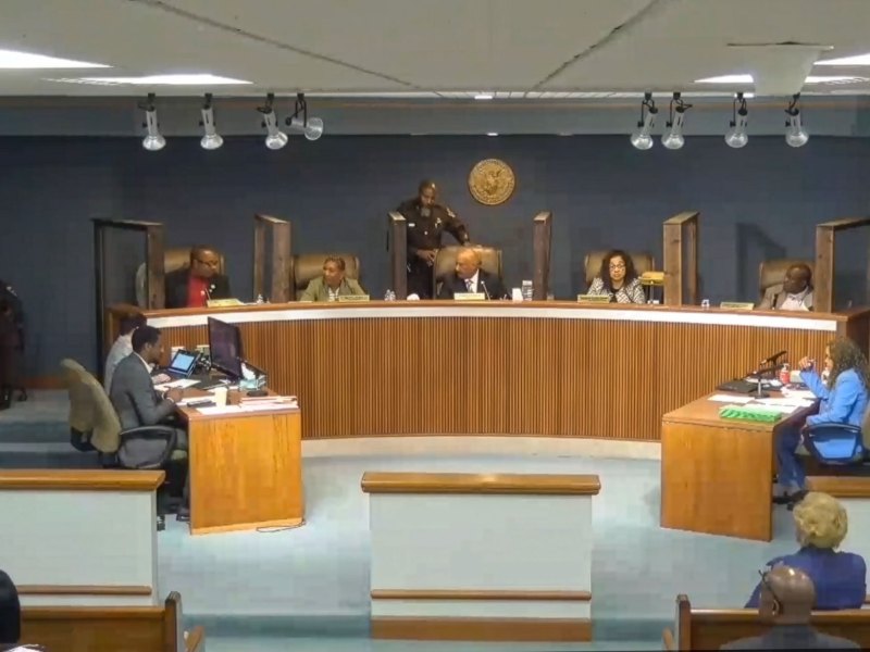 A man in a brown sheriff uniform walks behind a desk that seats two women and three men, facing two occupied desks and an audience.