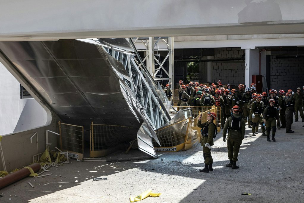A group of people wearing uniforms gather near a large steel, collapsed structure that appears to be part of a bridge