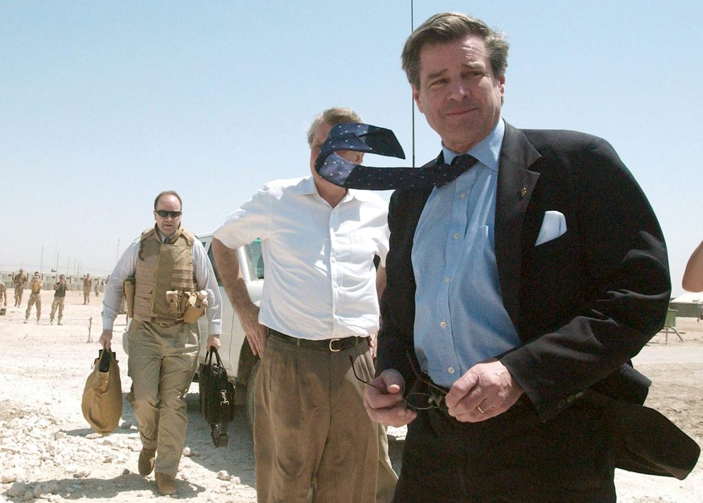 A man in a suit walking with the wind blowing his tie upward. A second man wearing a white shirt and a third man wearing a tactical vest are behind him