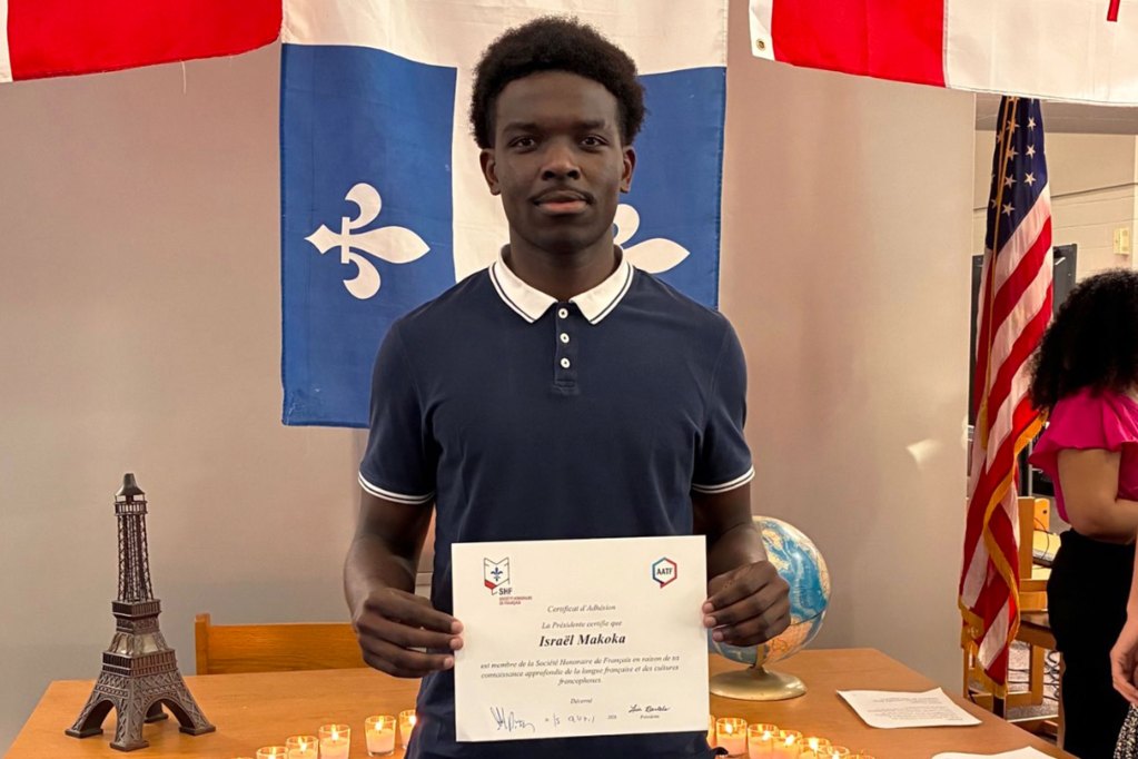 A teen stands in front of French flags with a certificate for Israel Makoka being in the French Honors Club