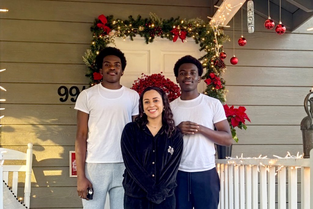 Two brothers stand behind a shorter woman outside of a house covered in green and red Christmas decorations