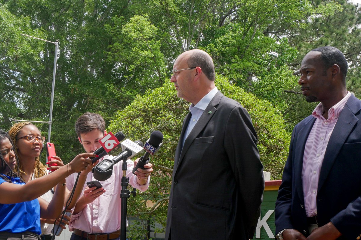 Two men stand in front of a group of three reporters, who are holding microphones and cell phones.