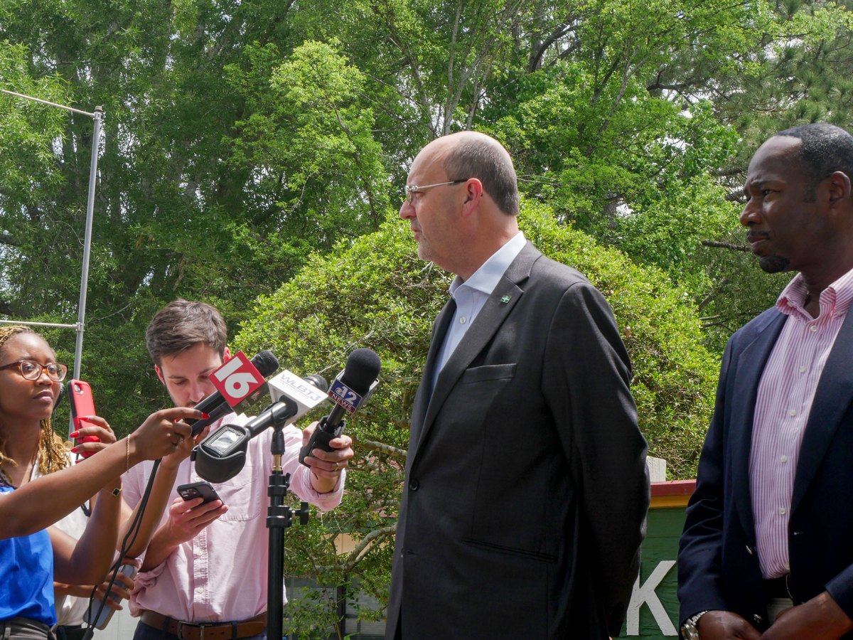 Two men stand in front of a group of three reporters, who are holding microphones and cell phones.