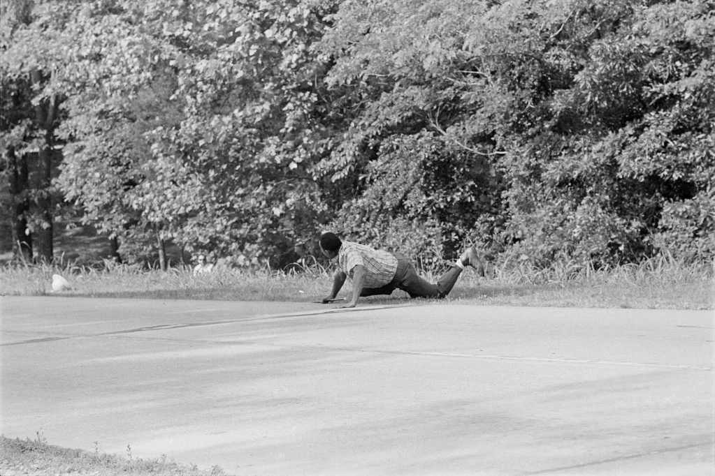 An older black and white photo of James Meredith crawling by the roadside, looking behind him at a person obscured by foliage