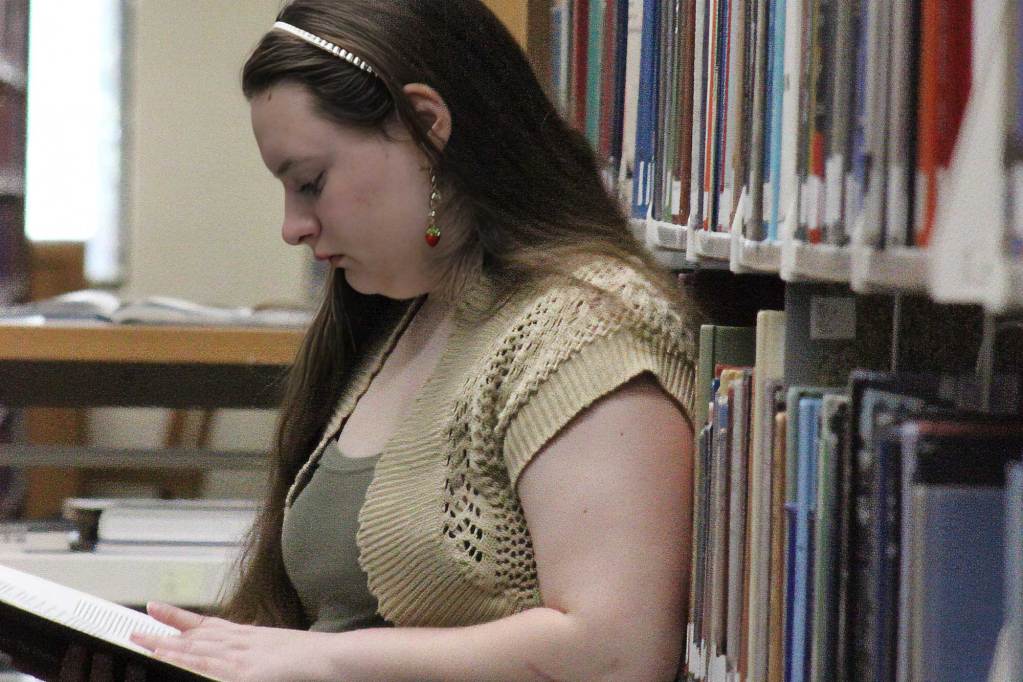 A woman leans against the shelves in a library and looks through an open book