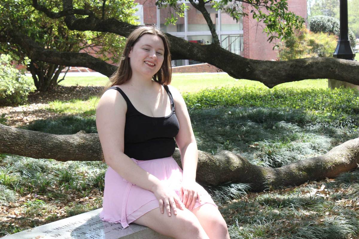 A woman in a black tank top and pink skirt sits on a bench beside a large tree