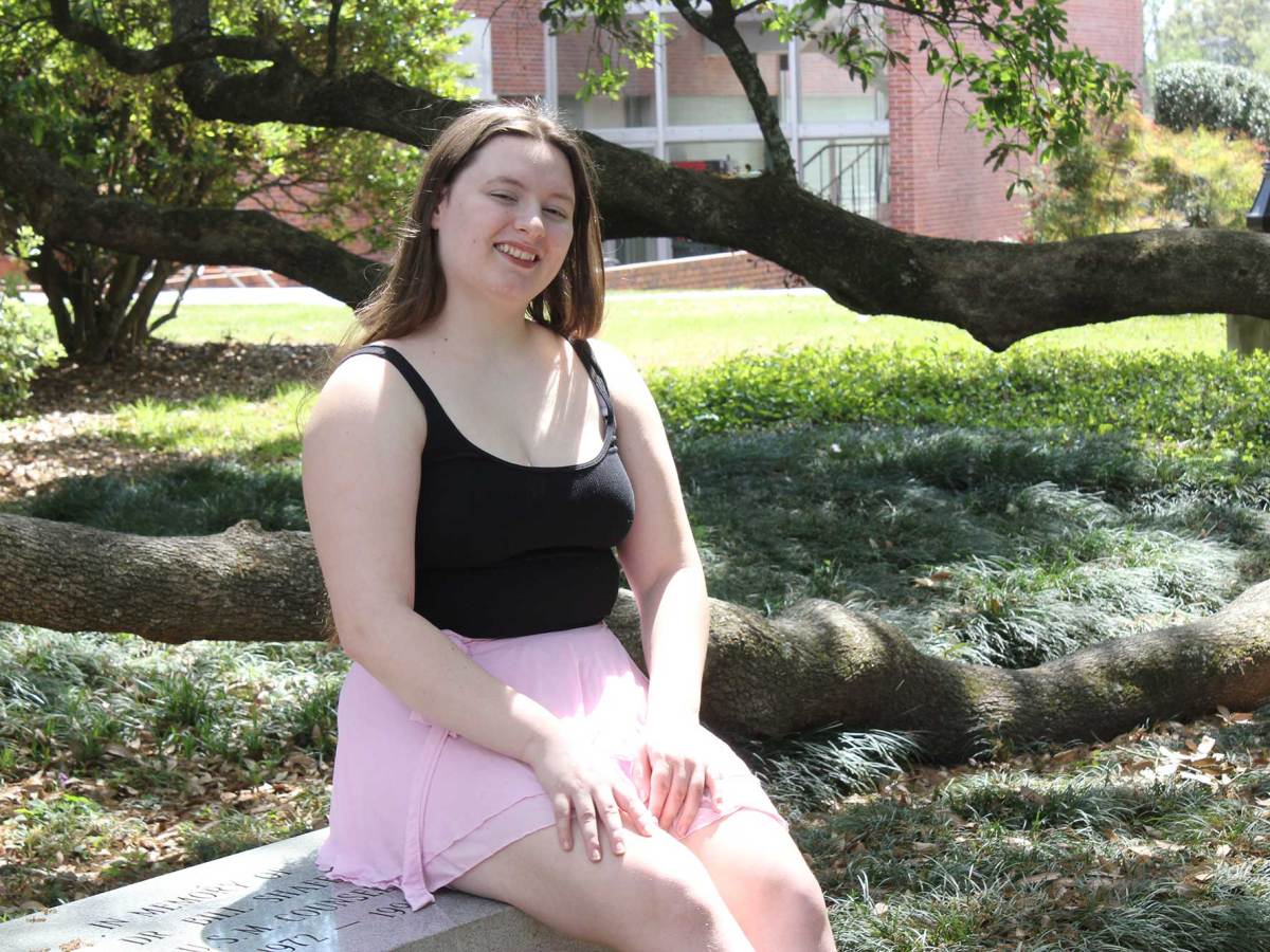 A woman in a black tank top and pink skirt sits on a bench beside a large tree