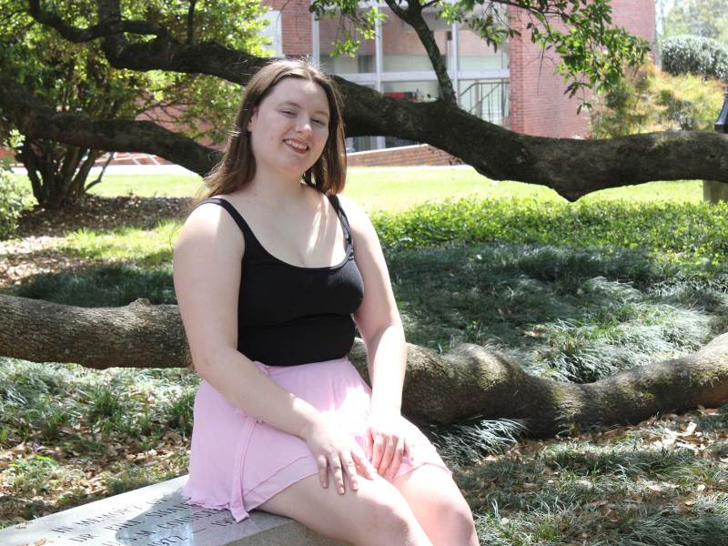 A woman in a black tank top and pink skirt sits on a bench beside a large tree