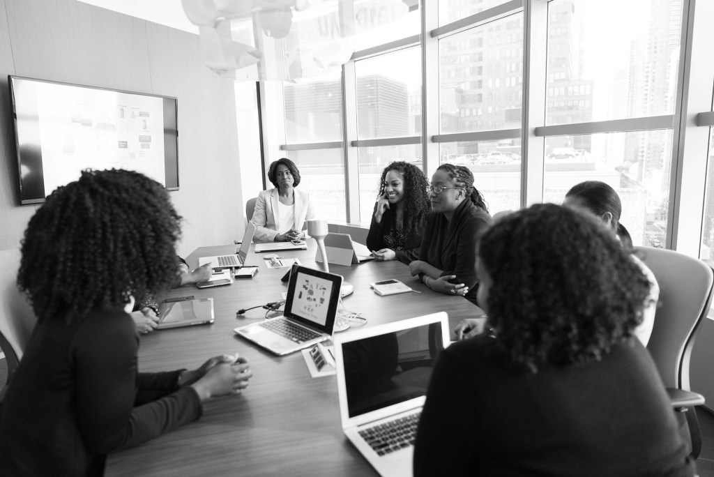 A black-and-white photo of several women seated at a conference table with laptops.