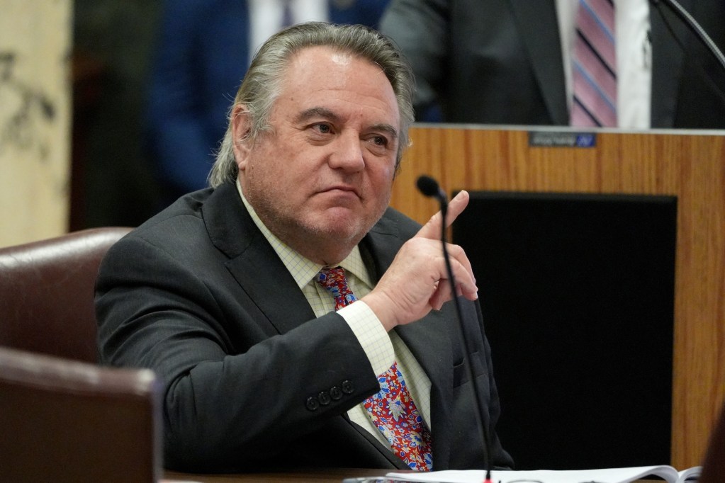 A seated an in a suit and red floral tie speaks with one hand pointing