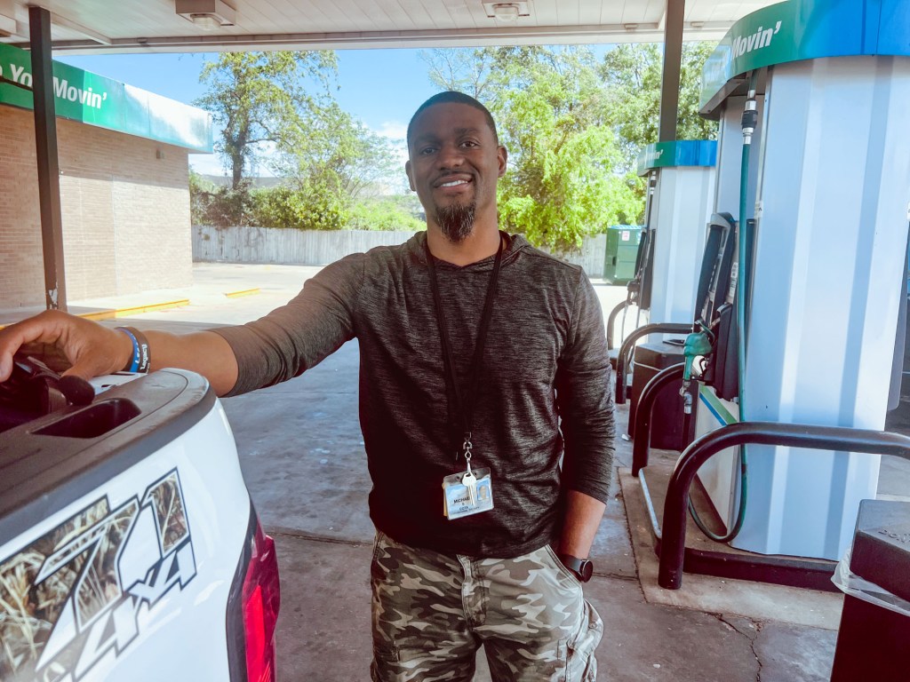 a black man smiles beside a white truck at a gas station