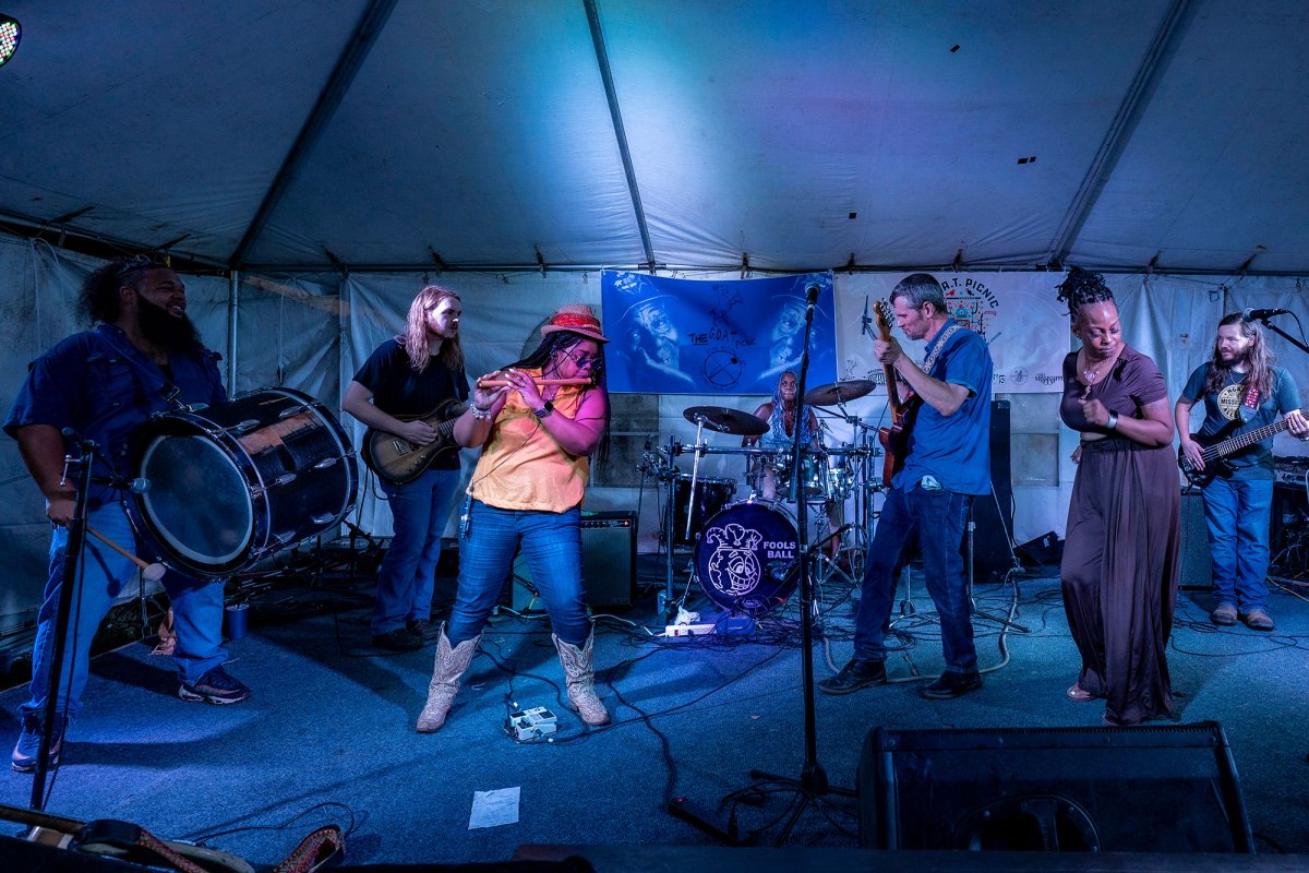 A six member band plays on stage with guitars, drums, and a flute