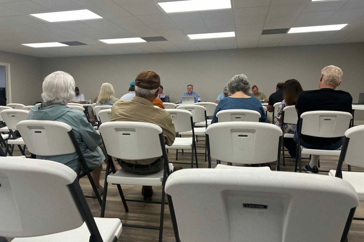 Thirteen people sit in white folding chairs in a room during a board meeting with water association officials.
