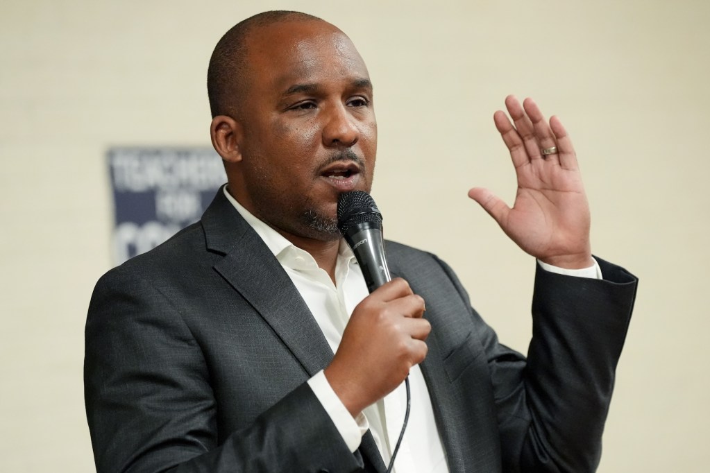 Scott Colom in a black suit and no tie speaks, mic in hand