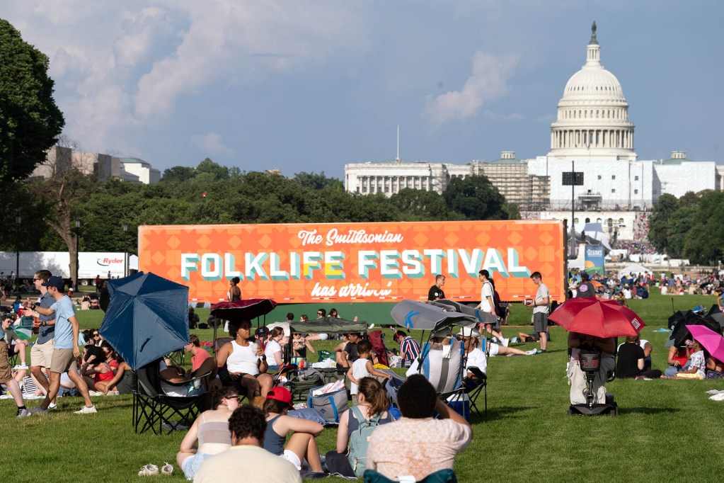 People on a green lawn with a large orange sign that reads 'The Smithsonian Folklife Festival has arrived'