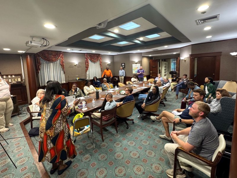 A large group of people seated at a conference table watching a presentation. Other people are seated near the table. 