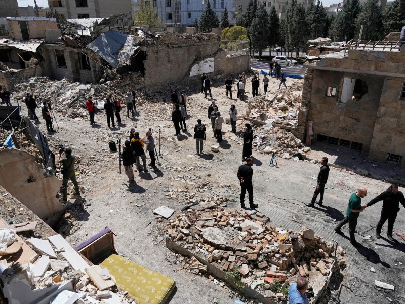 A group of people surveying a block of damaged buildings with rubble covering the ground.