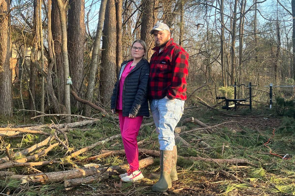 A couple wear jackets and stand in a yard filled with fallen tree debris
